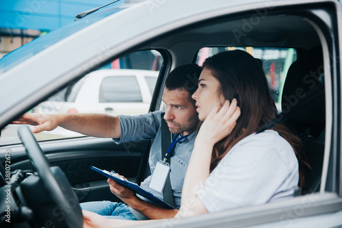 Male auto instructor explains driving rules to female student. Caucasian man points to the road and holds his paper. Young woman fixes her hair and listens to comments about her mistakes.