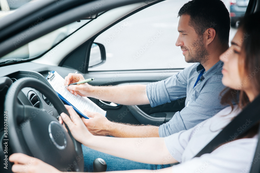 Satisfied male driving instructor notes results of test in his paper. Smiled man sitting on front passenger seat. Blurred brunette female student holding wheel and concentrated on the road. Satisfied male driving instructor notes results of test in his paper. Smiled man sitting on front passenger seat. Blurred brunette female student holding wheel and concentrated on the road.