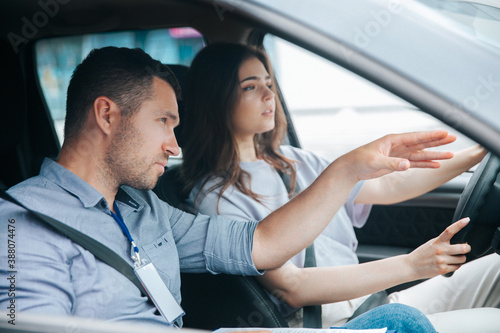 Male instructor showing something by his hand on the road and giving advice. Attractive woman listening her teacher, looking ahead with concentration and holding wheel. © Vlad
