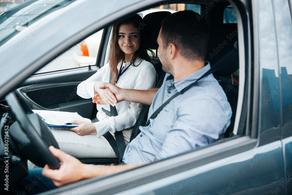 Female auto instructor shaking hands with male student. Attractive woman congratulates with successful driving license exam. Man in blue shirt holding wheel by his hand and looking at woman.