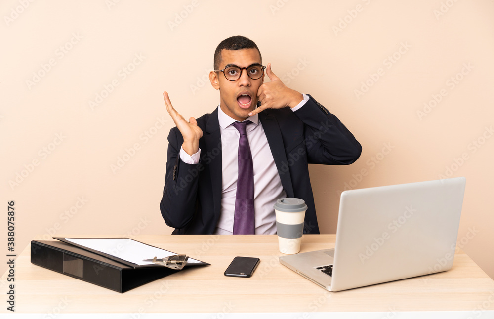 Young business man in his office with a laptop and other documents making phone gesture and doubting Young business man in his office with a laptop and other documents making phone gesture and doubting