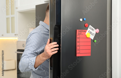 Man opening refrigerator door with shopping list, note and magnets in kitchen photo