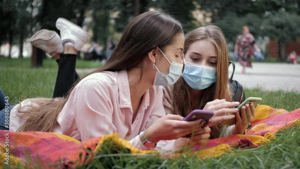Young women in medical masks lie in the park on the grass and using smartphone or cell phone. Tapping, scrolling, watching video, content, blogs. Young women in medical masks lie in the park on the grass and using smartphone or cell phone. Tapping, scrolling, watching video, content, blogs.
