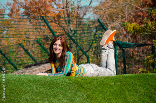 Cute redhaire woman in jeans, orange sneakers, rainbow blouse, smile happy, lay on stomach on green grass on hill in park photo