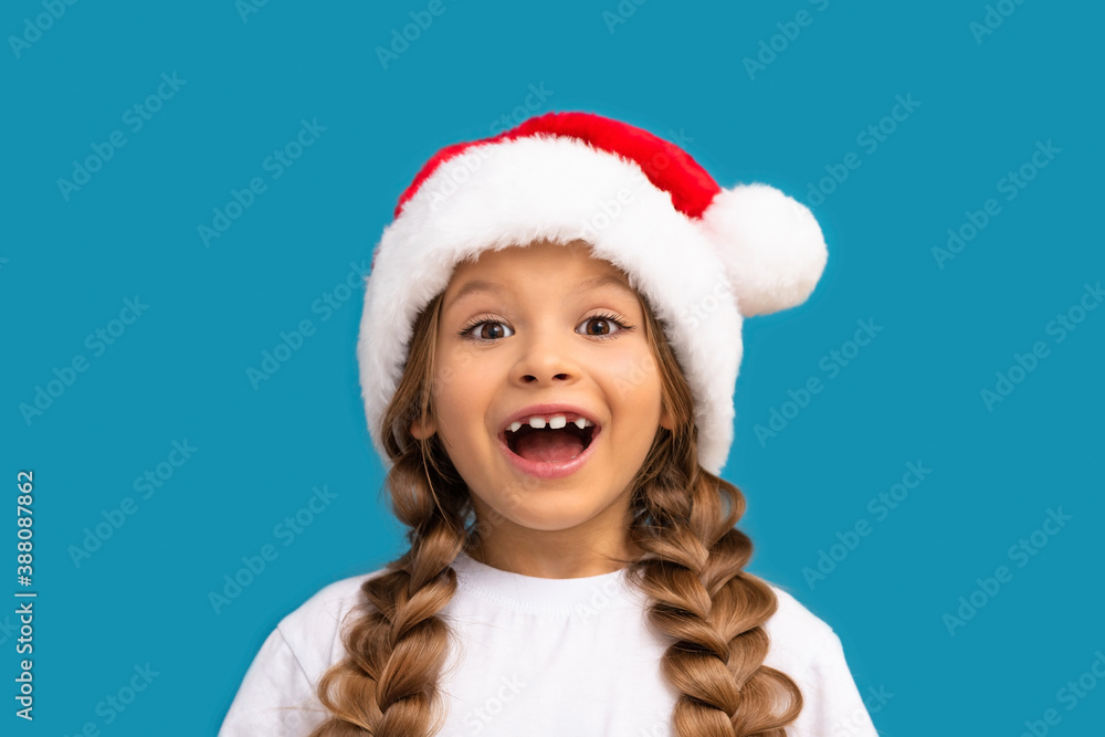 A little girl in a Christmas hat poses on a blue background. A little girl in a Christmas hat poses on a blue background.