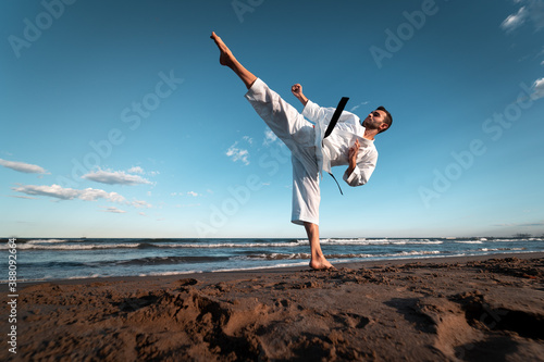 Black belt karate fighter giving a high kick on the beach at sunset photo