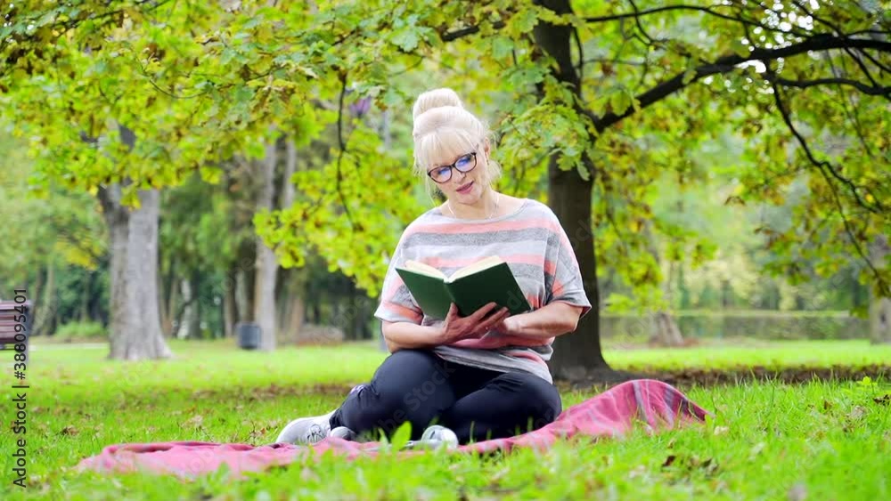 Portrait Happy Senior Woman Sitting in the Park on the Lawn Reading a Book holding in hands. Mature Female Relax and Rest in Nature Outdoors of the Countryside on grass. concept: Leisure in Old Age. Portrait Happy Senior Woman Sitting in the Park on the Lawn Reading a Book holding in hands. Mature Female Relax and Rest in Nature Outdoors of the Countryside on grass. concept: Leisure in Old Age.
