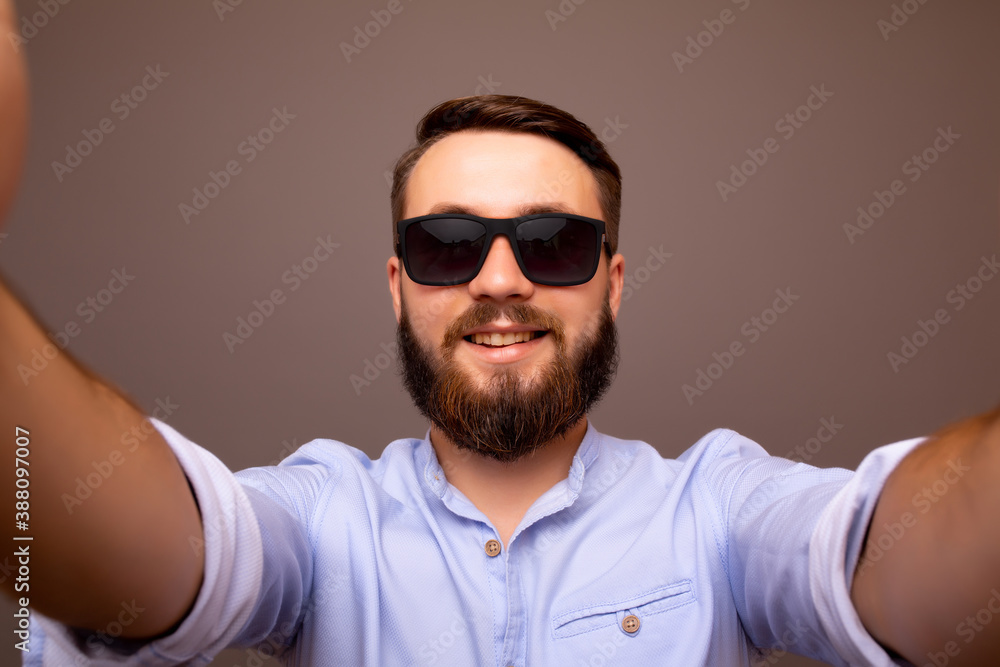 Obraz premium Close up of cheerful young bearded man in classic blue shirt and sunglasses make selfie isolated on grey background. 