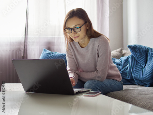 Smiling woman working on a laptop. Young, satisfied girl with glasses sitting on the couch in front of the laptop. There's a smartphone on the table.