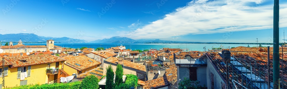 Panorama of Desenzano del Garda town with red tiled roof buildings, Garda Lake water, Monte Baldo mountain range, Sirmione peninsula, Lombardy, Northern Italy. Aerial panoramic view of Desenzano