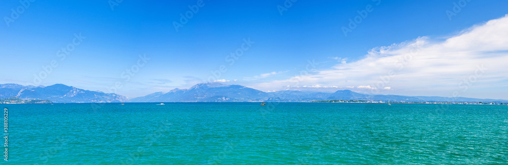 Panorama of Garda Lake azure turquoise water with view of Monte Baldo mountain range and Sirmione peninsula, blue sky white clouds background, Desenzano del Garda town, Lombardy, Northern Italy