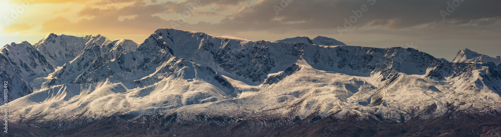 Scenic panoramic aerial view of snowy mountain peaks and slopes of North Chuyskiy ridge at sunset. Beautiful cloudy orange sky as a background. Altai mountains, Siberia, Russia