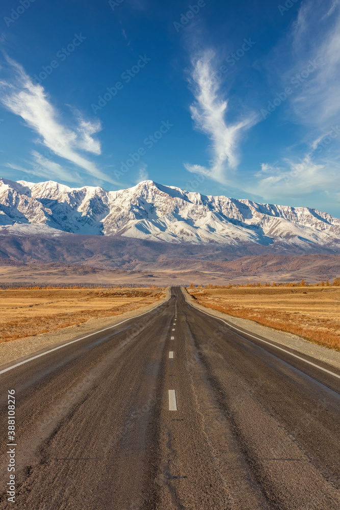 Portrait size shot of a straight empty highway leading to the snowy peaks of The Kuray mountain range. Beautiful blue cloudy sky as a background. Altai mountains, Siberia, Russia
