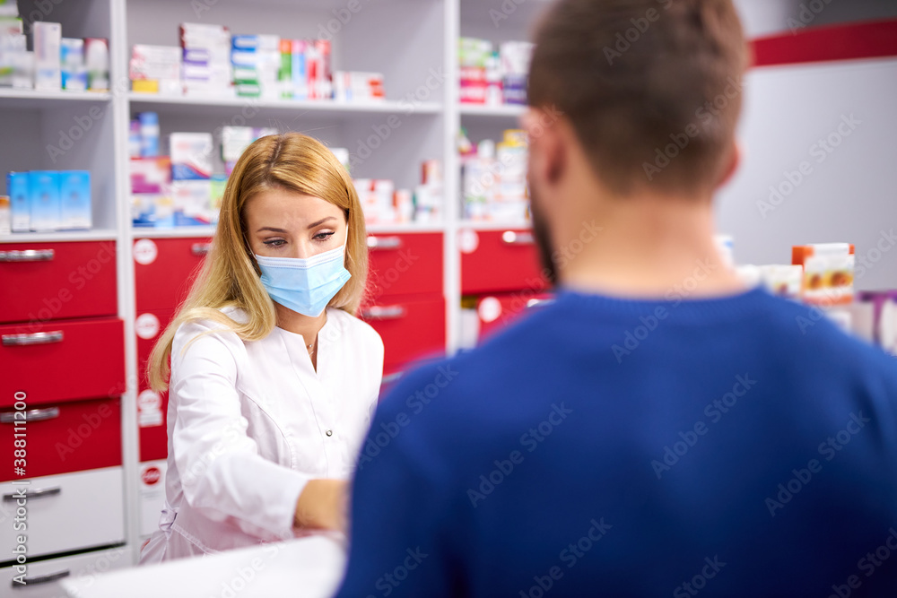 Obraz premium young female apothecary in protective medical mask and young caucasian man customer buying drug at drugstore. medicine, consumerism concept. during coronavirus epidemic