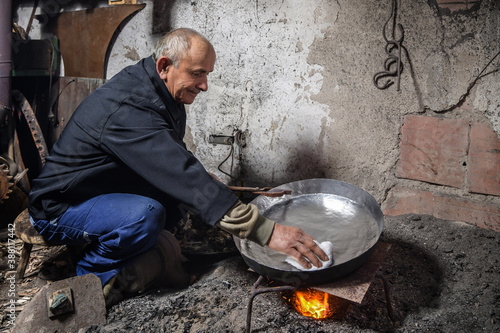 Elderly man in his workshop plates the copper bowl with tin. Tinsmith covering the copper object with tin over fire photo