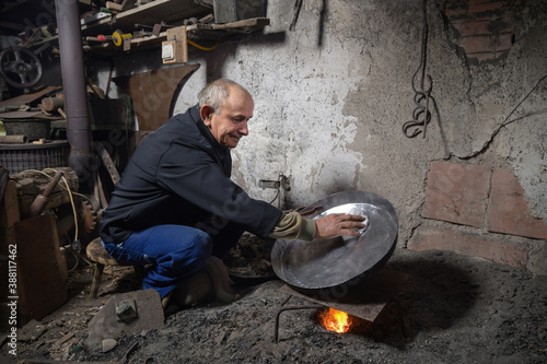 Elderly man in his workshop plates the copper bowl with tin. Tinsmith covering the copper object with tin over fire photo