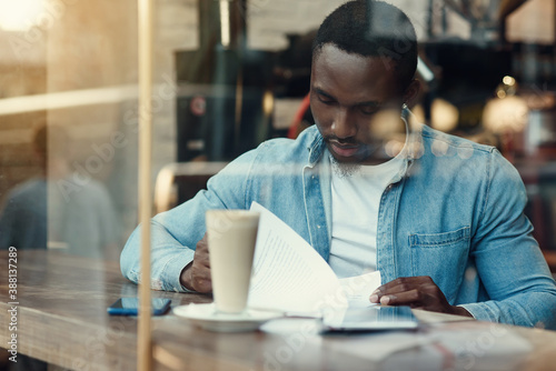 Concentrated black male entrepreneur reads paper documents sitting in cafe with coffee near window.