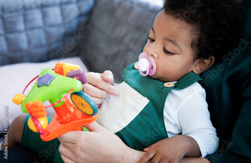 African American baby boy with pacifier is interesting his favorite toys while sit on mother's lap in living room. Portrait of a cute little happy black baby boy with mother at home, Family concept.