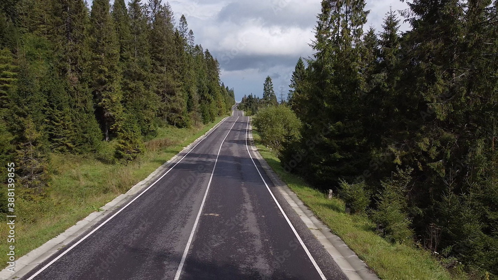 Naklejka premium Aerial low air flight over mountain perfect asphalt highway road and meadow at sunny summer morning. Near green trees. Fast horizontal movement with sun rays.