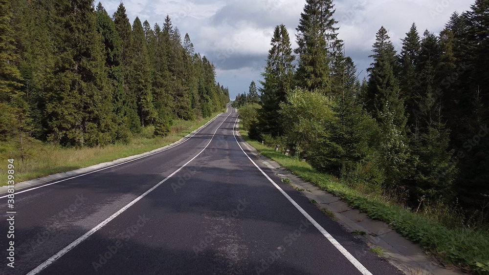 Aerial low air flight over mountain perfect asphalt highway road and meadow at sunny summer morning. Near green trees. Fast horizontal movement with sun rays.