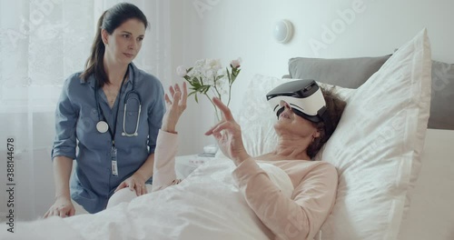 Female Doctor Checking on Elderly Patient Lying in Hospital Bed Doing Therapy Via VR Technology. Friendly Doctor Helping Senior Woman Receive Therapy Using Virtual Reality Headset.