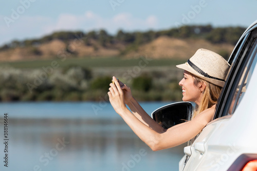 Young woman leaning on car window while taking photo with phone sitting in car photo