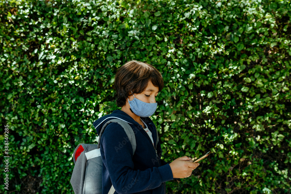 Schoolboy wearing mask using smart phone while standing against plants Schoolboy wearing mask using smart phone while standing against plants