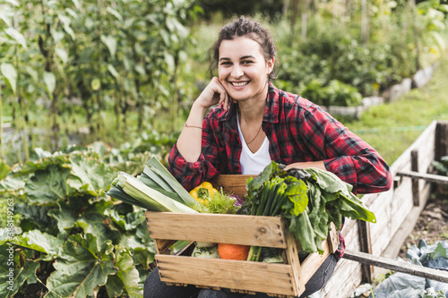 Fototapeta Smiling young woman sitting with vegetables in wooden crate at community garden