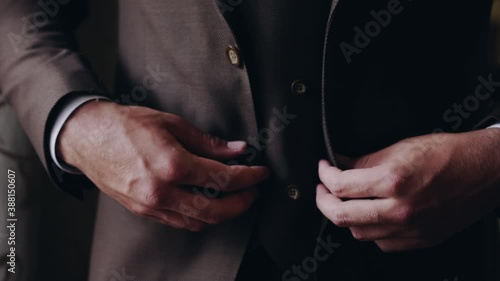 A well-groomed neat caucasian man elegantly fastens a button on a brown jacket worn over a vest. Close-up, light falling from the side