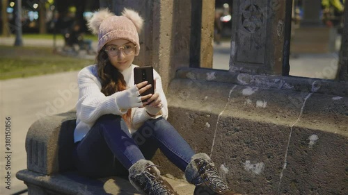 niña bonita,alegre vestida con ropa de invierno sentada tomándose fotos y mandando mensajes en el celular al aire libre photo