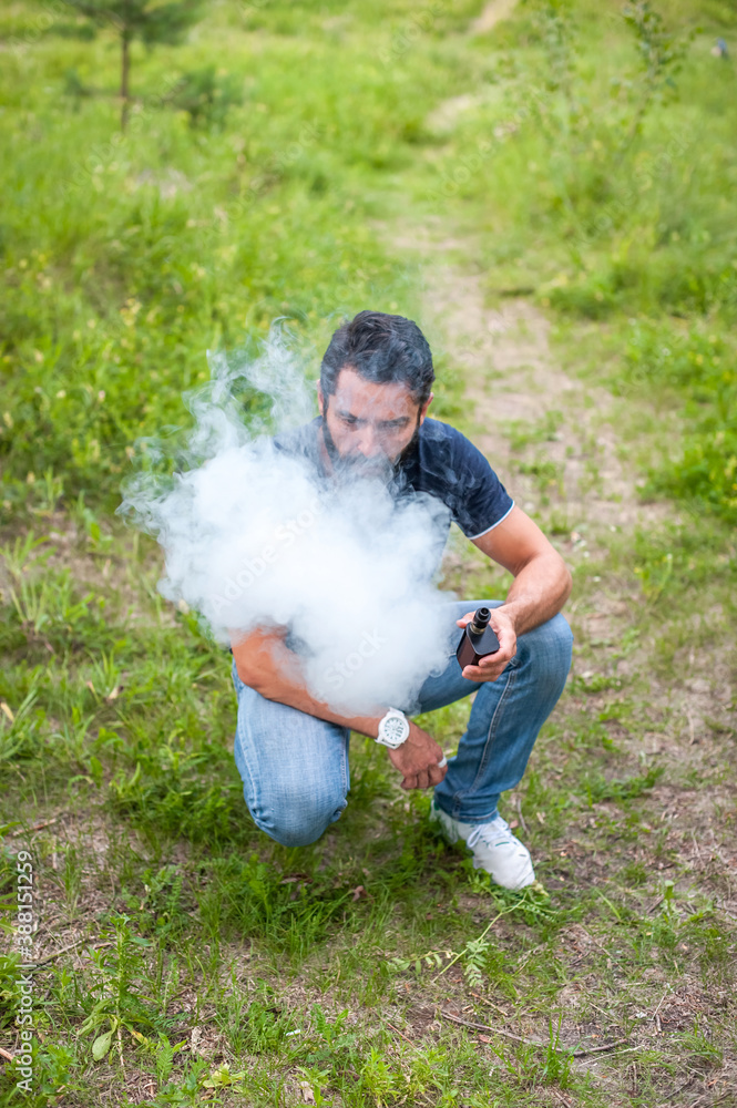 Bearded vape man is resting on the grass and vaping and letting off steam from an e-cigarette. Modern ways of quitting tobacco. Bearded vape man is resting on the grass and vaping and letting off steam from an e-cigarette. Modern ways of quitting tobacco.
