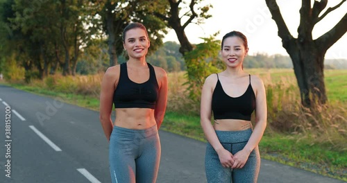 Mixed-races young beautiful women athletes standing on road after training and smilinf to camera. Joyful Caucasian and Asian sporty females outdoors in good mood. Sport concept photo