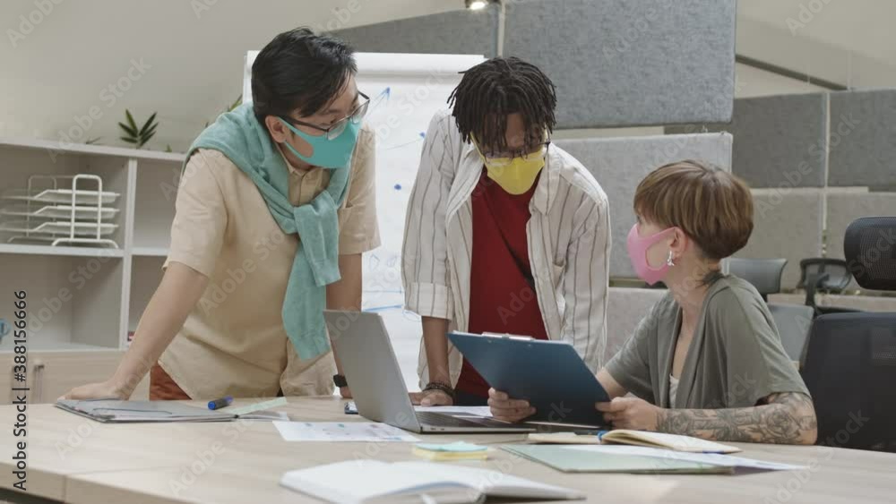 Medium shot of three young multiethnic coworkers wearing masks gathered together in office and having conversation concerning charts. Then they looking at camera Medium shot of three young multiethnic coworkers wearing masks gathered together in office and having conversation concerning charts. Then they looking at camera
