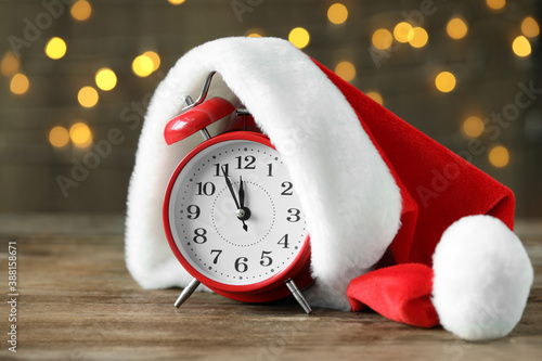 Alarm clock with Santa hat on wooden table against blurred Christmas lights. New Year countdown