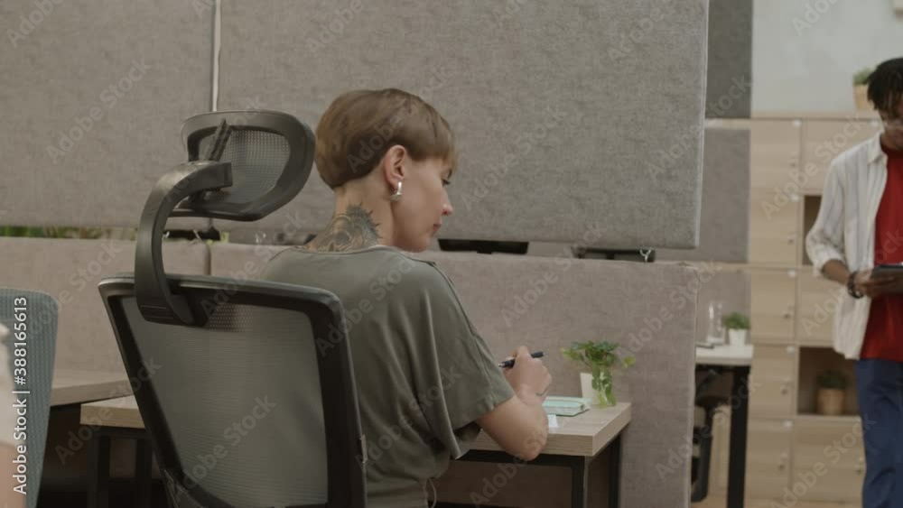 Panning of young pretty Caucasian woman with short haircut wearing casual clothes sitting at desk in office, working then turning around and looking at camera Panning of young pretty Caucasian woman with short haircut wearing casual clothes sitting at desk in office, working then turning around and looking at camera