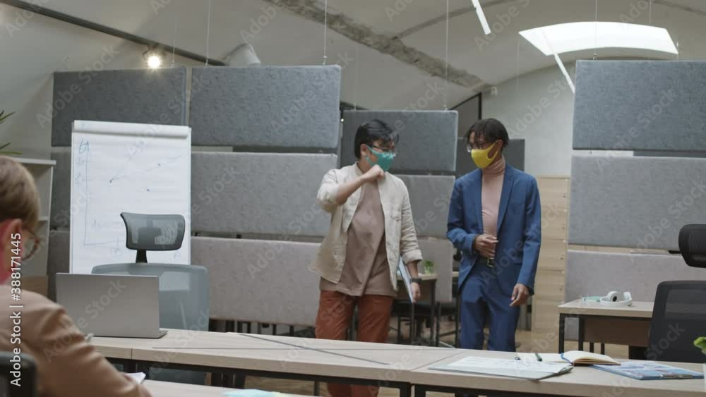 Medium shot of two diverse young men wearing masks coming in modern office, greeting each other and sitting down at workplaces. Caucasian woman doing paperwork Medium shot of two diverse young men wearing masks coming in modern office, greeting each other and sitting down at workplaces. Caucasian woman doing paperwork