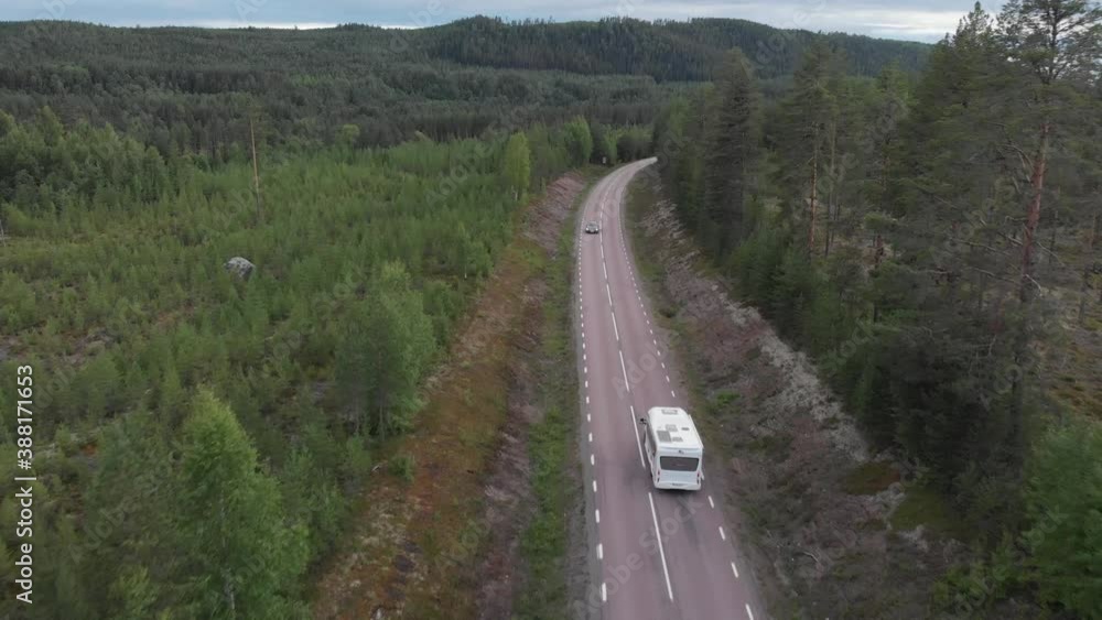 Aerial tracking shot behind camper van crossing along forest road, surrounded by high pine trees, in mountainous environment Aerial tracking shot behind camper van crossing along forest road, surrounded by high pine trees, in mountainous environment