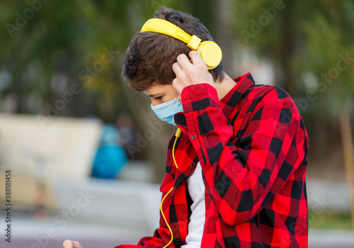 Close up cute boy with headphones and protective mask on face. Teenager in red check flannel shirt wearing medical mask to protect from corona virus, illness, infection. Quarantine on the city streets