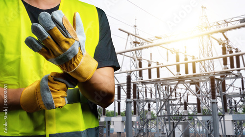 An electrician wearing a black T-shirt and personal protective equipment. Are wearing leather gloves to prepare for work. There is a high voltage power station in the background.