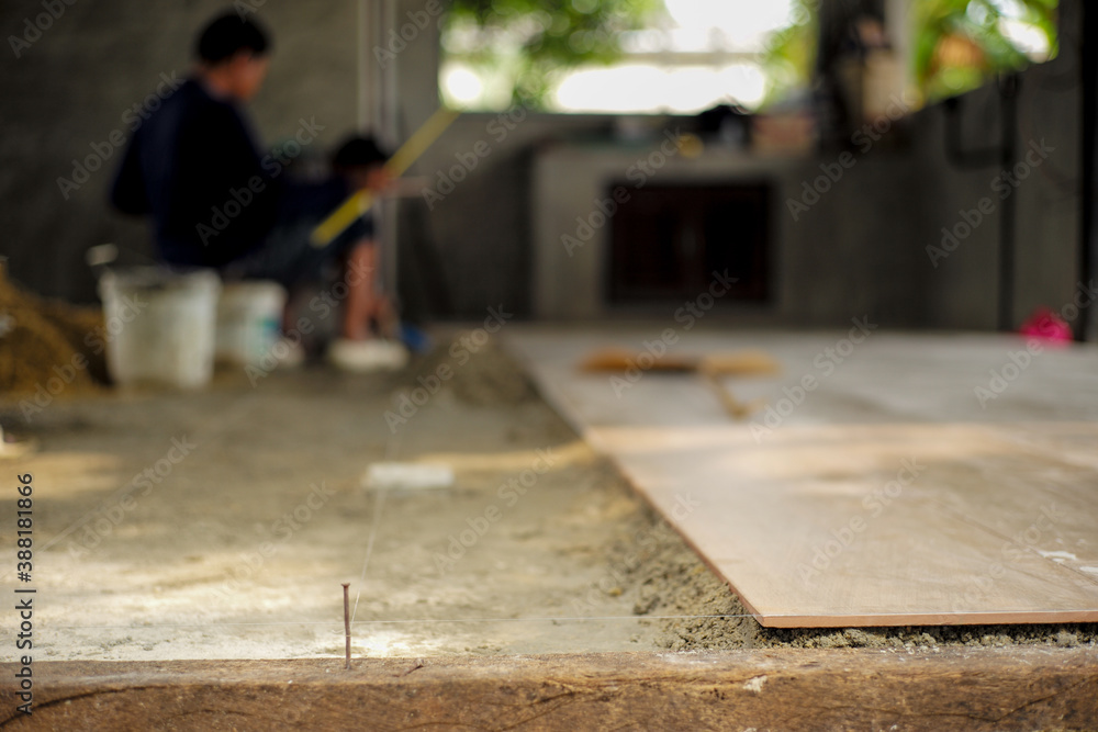 Closeup floor tiles on the mixed cement with nylon for levelling in the house under construction with blurred workers in background Closeup floor tiles on the mixed cement with nylon for levelling in the house under construction with blurred workers in background