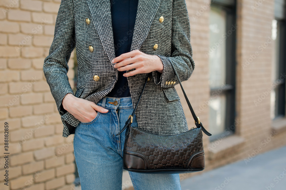 Fototapeta premium Young fashionable woman wearing gray tweed jacket with golden buttons, blue jeans, sweater and holding black leather baguette handbag. Street style.