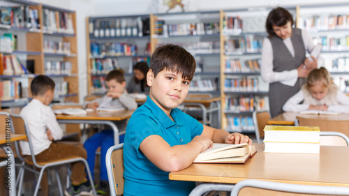 Portrait of smiling intelligent preteen boy reading in school library on background with other students and teacher 
