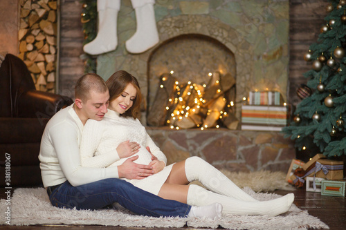 Pair of future parents near christmas fireplace. Pregnant woman hugging her huband & belly with hands in Christmas home. Family at Cozy home. Romantic parents waiting for baby. Casual clothes outfit. photo