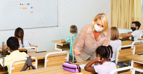 Teacher woman in protective mask helping schoolkid during lesson in schoolroom