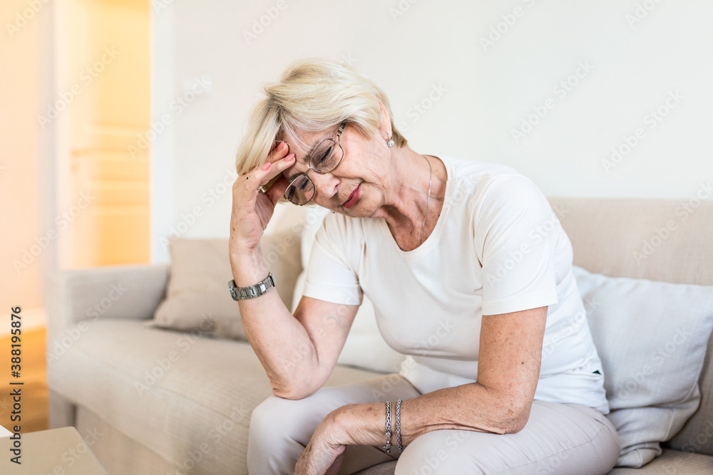 Mature woman sitting on a white sofa in a home touching her head with her hand while having a headache pain and feeling unwell Mature woman sitting on a white sofa in a home touching her head with her hand while having a headache pain and feeling unwell