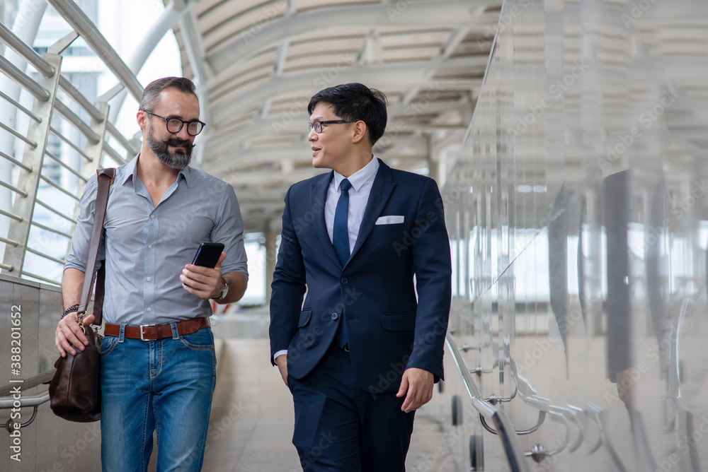 Two businessman walking together on office district and discussing business project with using smartphone. Caucasian beard man with Asian man office worker colleague talking about working plan Two businessman walking together on office district and discussing business project with using smartphone. Caucasian beard man with Asian man office worker colleague talking about working plan