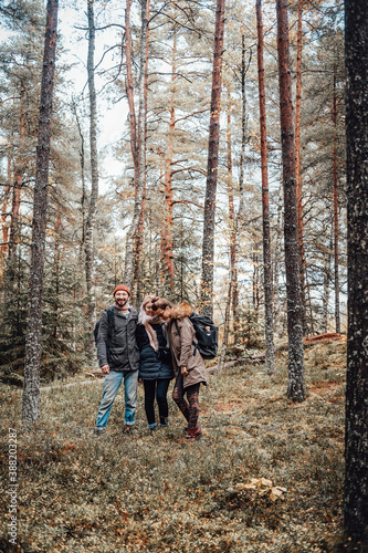 Cheerful and happy group of three tourists pose in autumn forest around trees and look at camera.