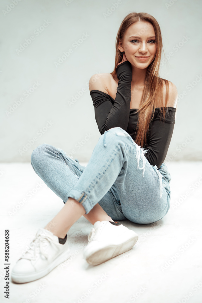 Attractive and smiley girl with brown hairs and slim figure in urban stylish clothing poses sitting on floor in white background. Attractive and smiley girl with brown hairs and slim figure in urban stylish clothing poses sitting on floor in white background.