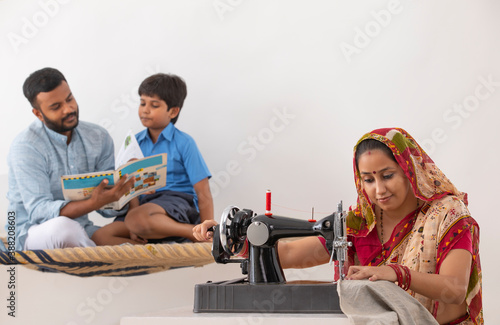 PORTRAIT OF A RURAL WOMAN SEWING CLOTHES WHILE HUSBAND TEACHING KID IN THE BACKGROUND 
