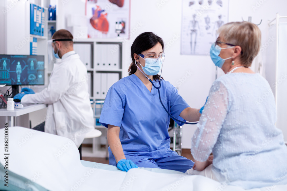 Obraz premium Nurse with face mask listening patient heart using stethoscope in hospital examination room wearing protection gear. Medical examination for infections, disease and diagnosis.